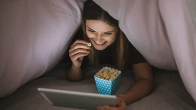 Jovem assiste a vídeos em um tablet sob as cobertas, com balde de pipoca