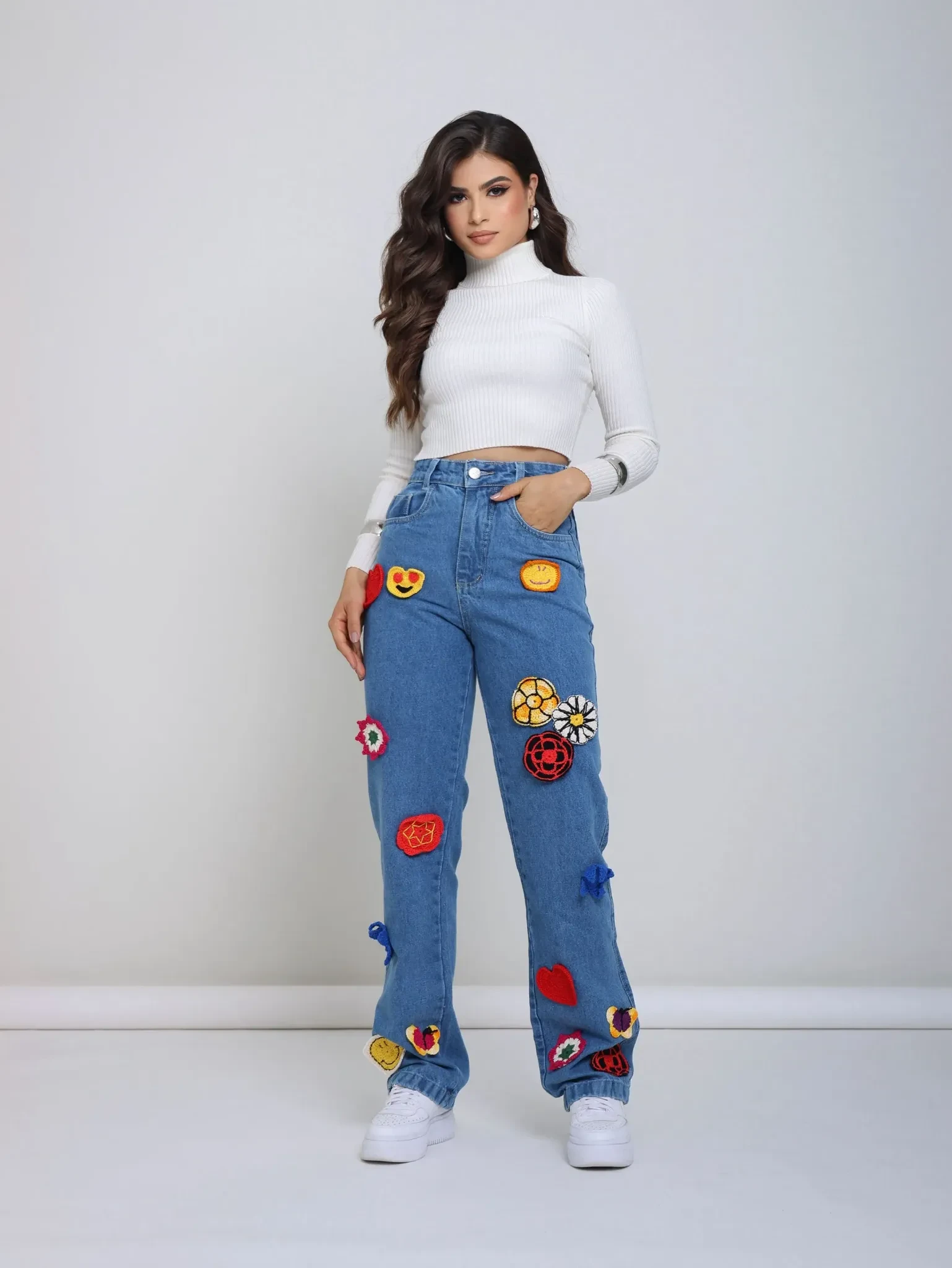 Calça jeans azul com diversos patches de crochê coloridos, combinada com blusa branca de manga longa.