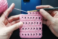Mãos crochetem uma capinha rosa para celular, com dispositivo azul ao fundo.