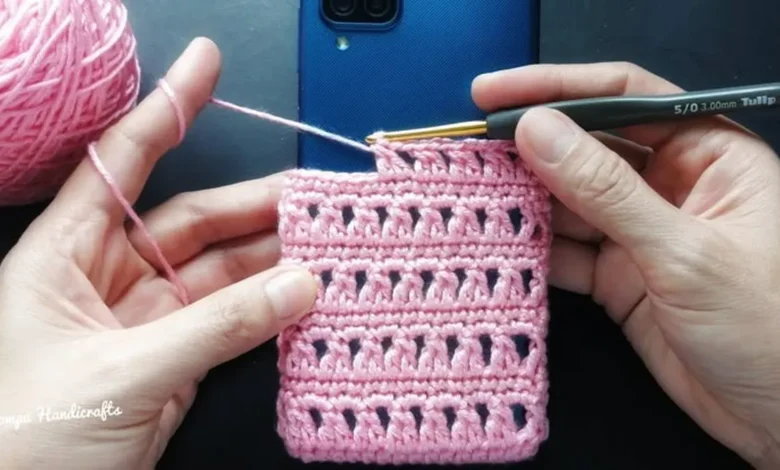Mãos crochetem uma capinha rosa para celular, com dispositivo azul ao fundo.