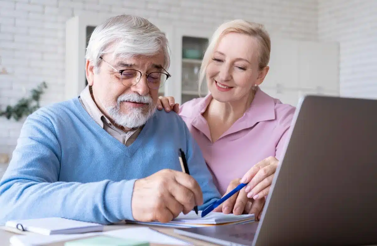 Casal idoso feliz planejando a aposentadoria juntos, com papéis e laptop à frente.