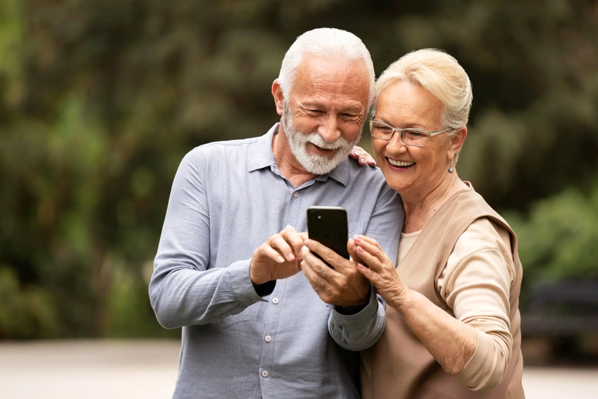 Casal idoso sorridente olhando juntos para o celular ao ar livre.
