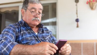 Homem idoso usando celular com capa roxa, sentado à mesa em ambiente doméstico, com expressão concentrada.