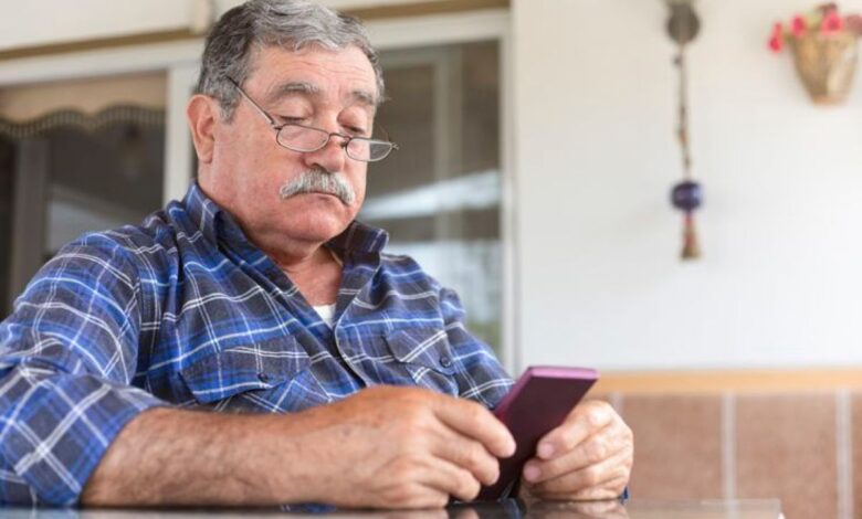 Homem idoso usando celular com capa roxa, sentado à mesa em ambiente doméstico, com expressão concentrada.