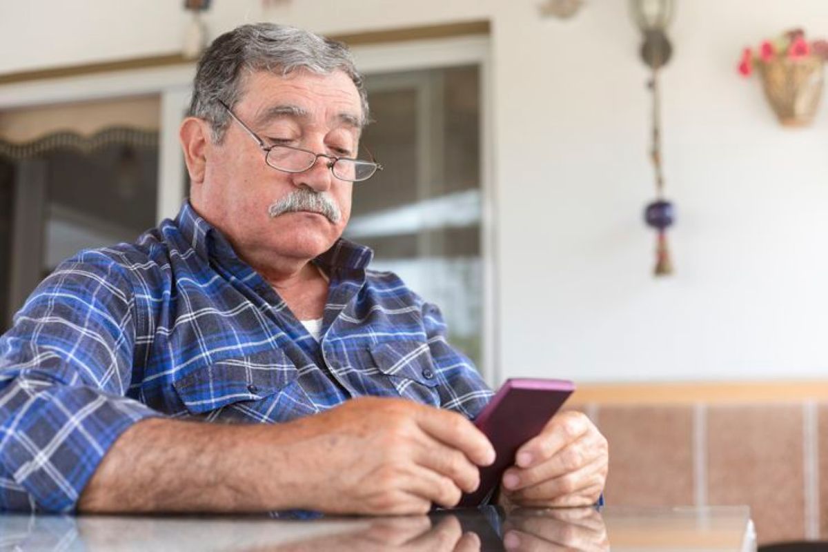 Homem idoso usando celular com capa roxa, sentado à mesa em ambiente doméstico, com expressão concentrada.