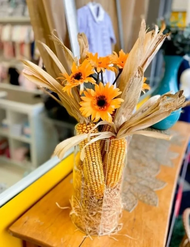 Arranjo decorativo de centro de mesa com espigas de milho seco e flores amarelas em jarra de vidro