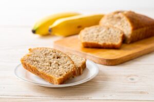 Fatias de pão de banana servidas em prato branco com bananas ao fundo sobre mesa de madeira clara