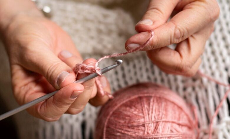 MĂŁos femininas fazendo crochĂȘ com fio rosa, representando o processo artesanal de criação de peças para cozinha