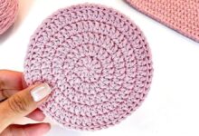 Disco circular de crochê rosa claro com pontos uniformes e formato plano, segurado por uma mão com unhas esmaltadas
