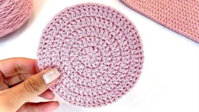 Disco circular de crochê rosa claro com pontos uniformes e formato plano, segurado por uma mão com unhas esmaltadas