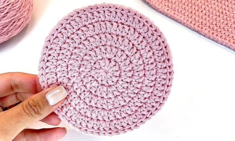 Disco circular de crochĂȘ rosa claro com pontos uniformes e formato plano, segurado por uma mĂŁo com unhas esmaltadas