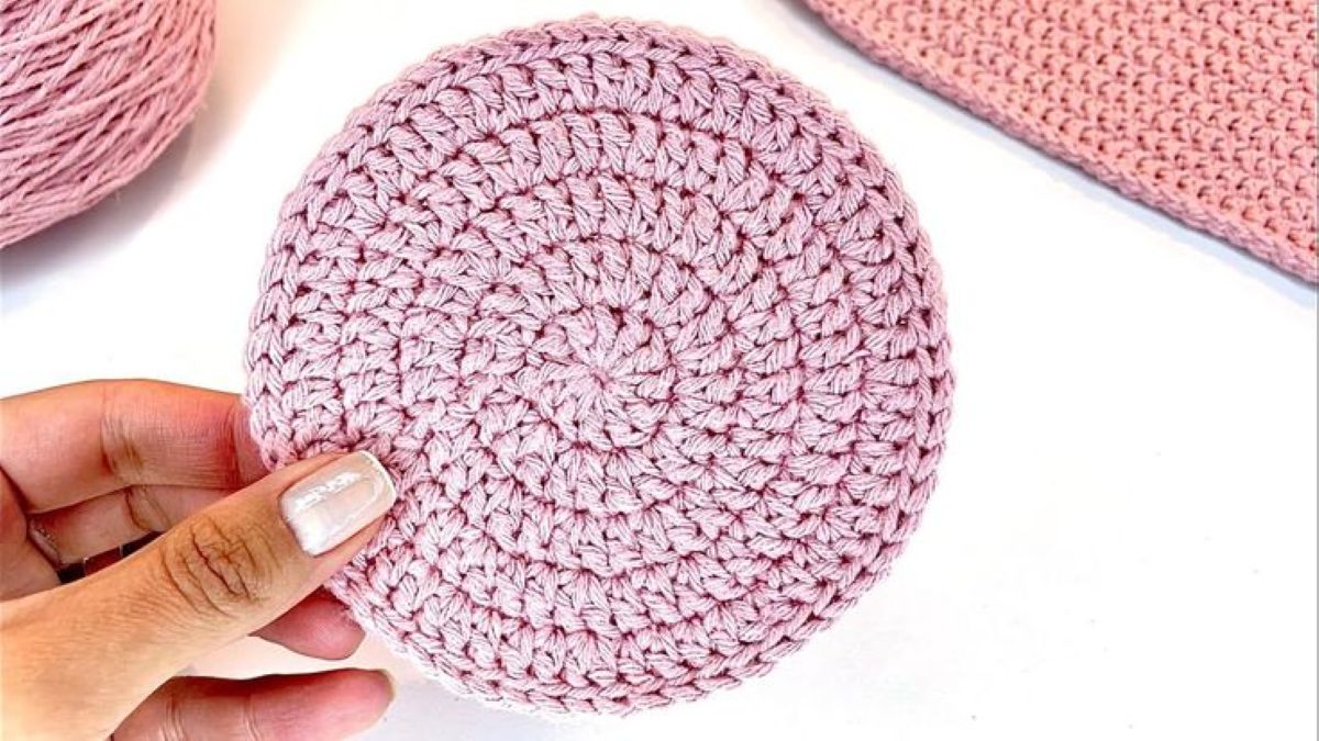 Disco circular de crochê rosa claro com pontos uniformes e formato plano, segurado por uma mão com unhas esmaltadas