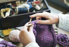 Mãos tricotando um cachecol roxo de crochê com fio grosso e agulhas, com materiais de costura ao fundo.