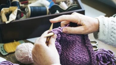 Mãos tricotando um cachecol roxo de crochê com fio grosso e agulhas, com materiais de costura ao fundo.