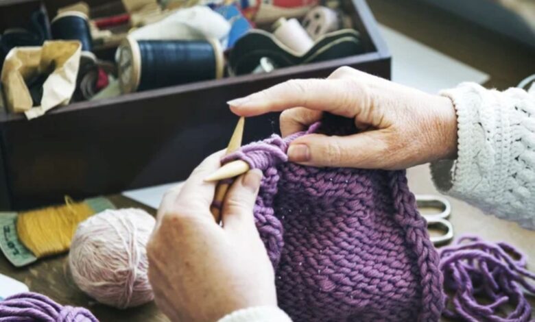 MĂŁos tricotando um cachecol roxo de crochĂȘ com fio grosso e agulhas, com materiais de costura ao fundo.