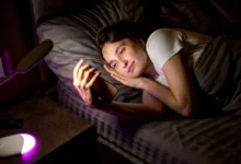 Pessoa sorridente deitada na cama, olhando para o celular à noite, transmitindo uma mensagem de boa noite cheia de carinho e aconchego