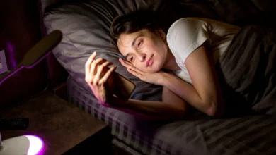Pessoa sorridente deitada na cama, olhando para o celular à noite, transmitindo uma mensagem de boa noite cheia de carinho e aconchego