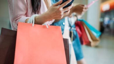 Consumidora verificando promoções no celular enquanto segura sacolas de compras
