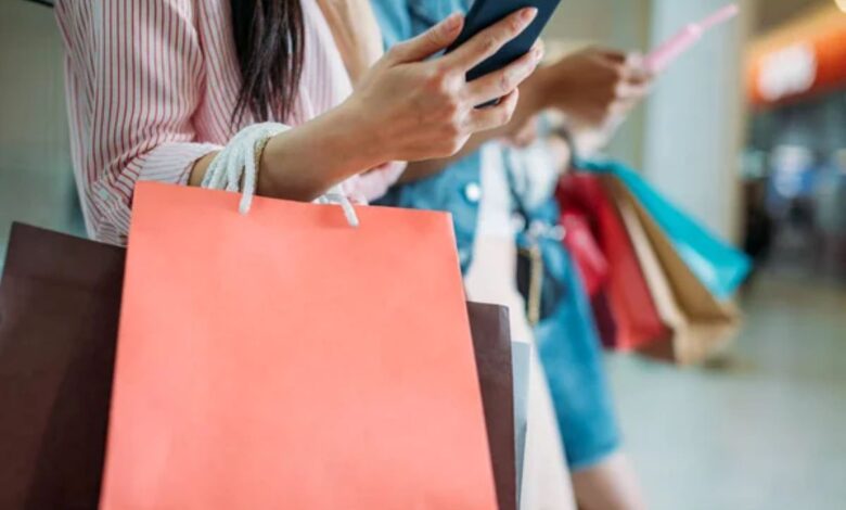 Consumidora verificando promoções no celular enquanto segura sacolas de compras