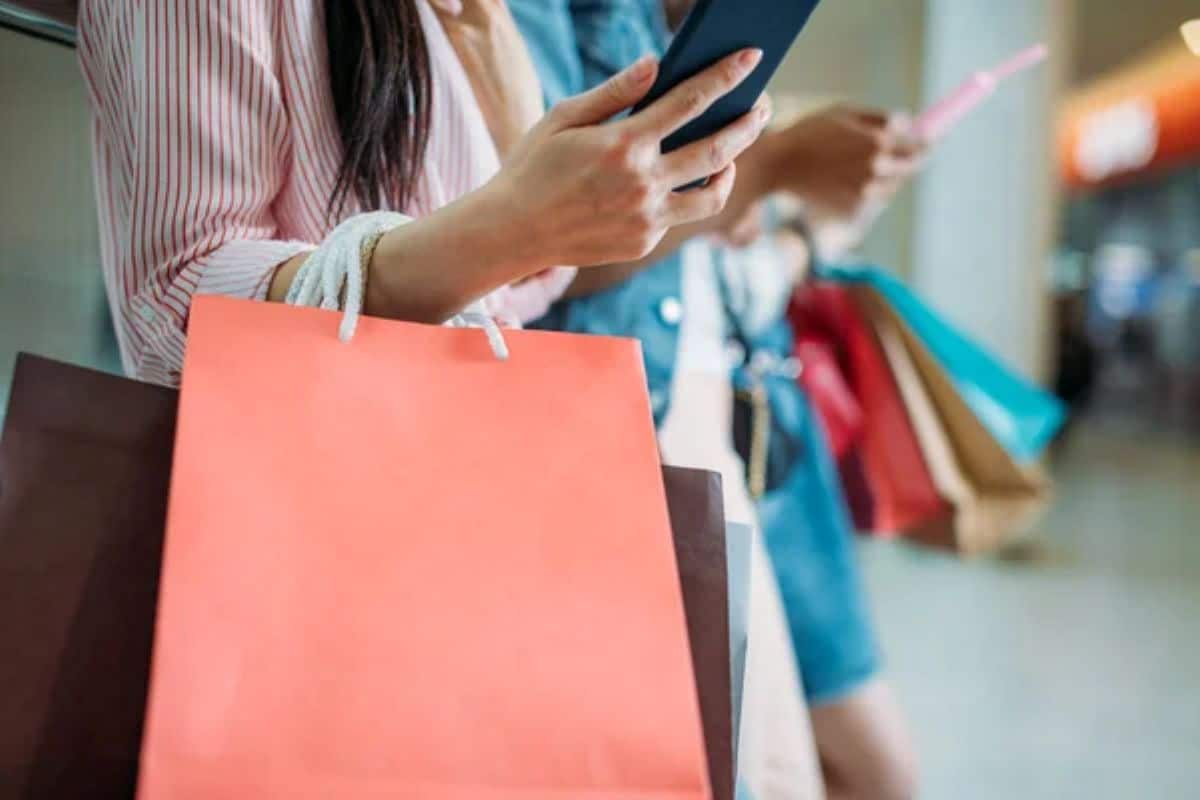 Consumidora verificando promoções no celular enquanto segura sacolas de compras