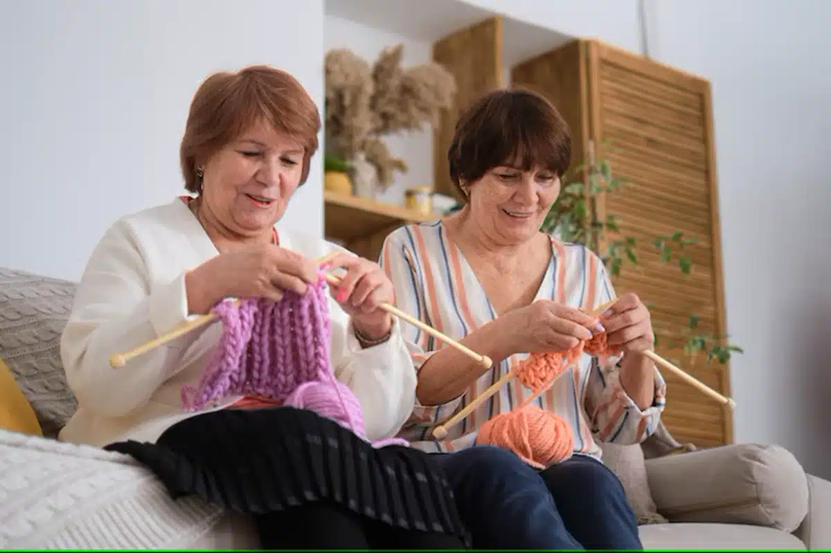 Duas mulheres idosas sorrindo enquanto fazem crochê com fios coloridos, demonstrando habilidade e concentração