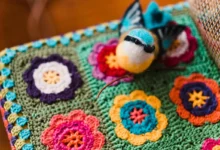 Peça de crochê colorida com flores e pássaro decorativo, mostrando uma embalagem criativa e artesanal para vendas.