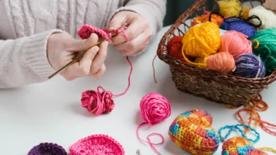 Mãos crocheteando com linha rosa, ao lado de uma cesta cheia de novelos coloridos e enfeites de crochê vibrantes em formato de coração.