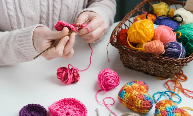 Mãos crocheteando com linha rosa, ao lado de uma cesta cheia de novelos coloridos e enfeites de crochê vibrantes em formato de coração.