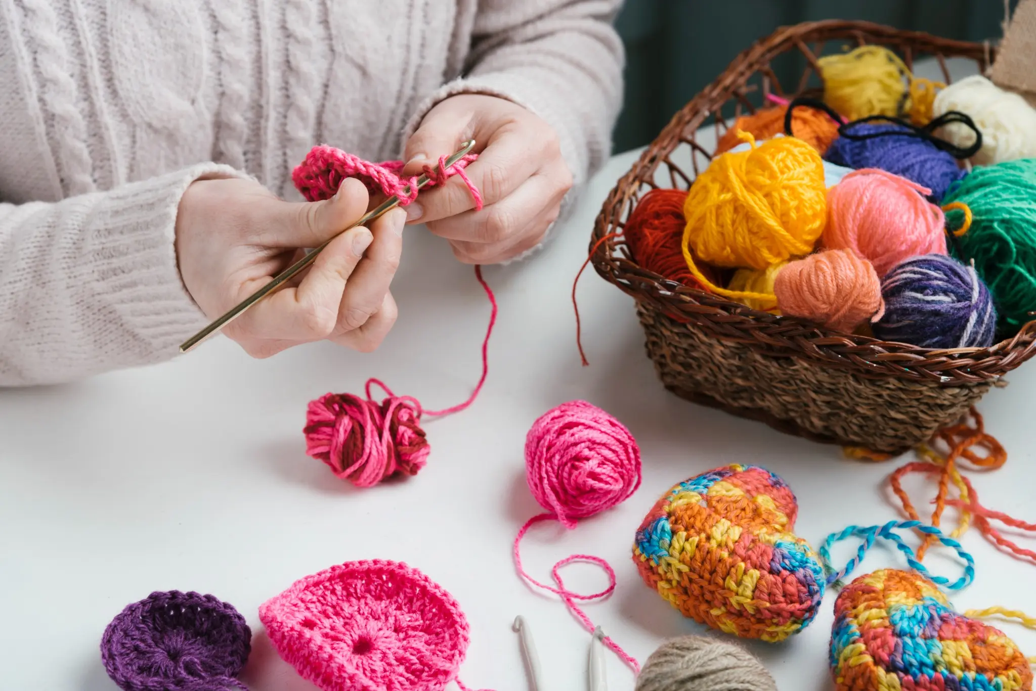 Mãos crocheteando com linha rosa, ao lado de uma cesta cheia de novelos coloridos e enfeites de crochê vibrantes em formato de coração.