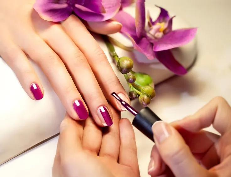 Mãos com unhas sendo esmaltadas em tom rosa tradicional, com flores roxas ao fundo.