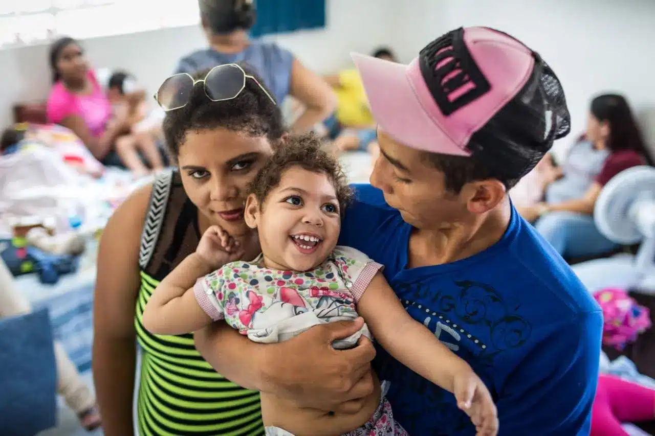 Criança sorridente com deficiência causada pelo vírus Zika sendo abraçada por adultos em ambiente comunitário.