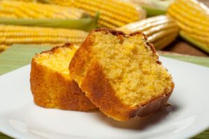 Fatias de bolo de milho servido em um prato branco, com espigas de milho ao fundo.