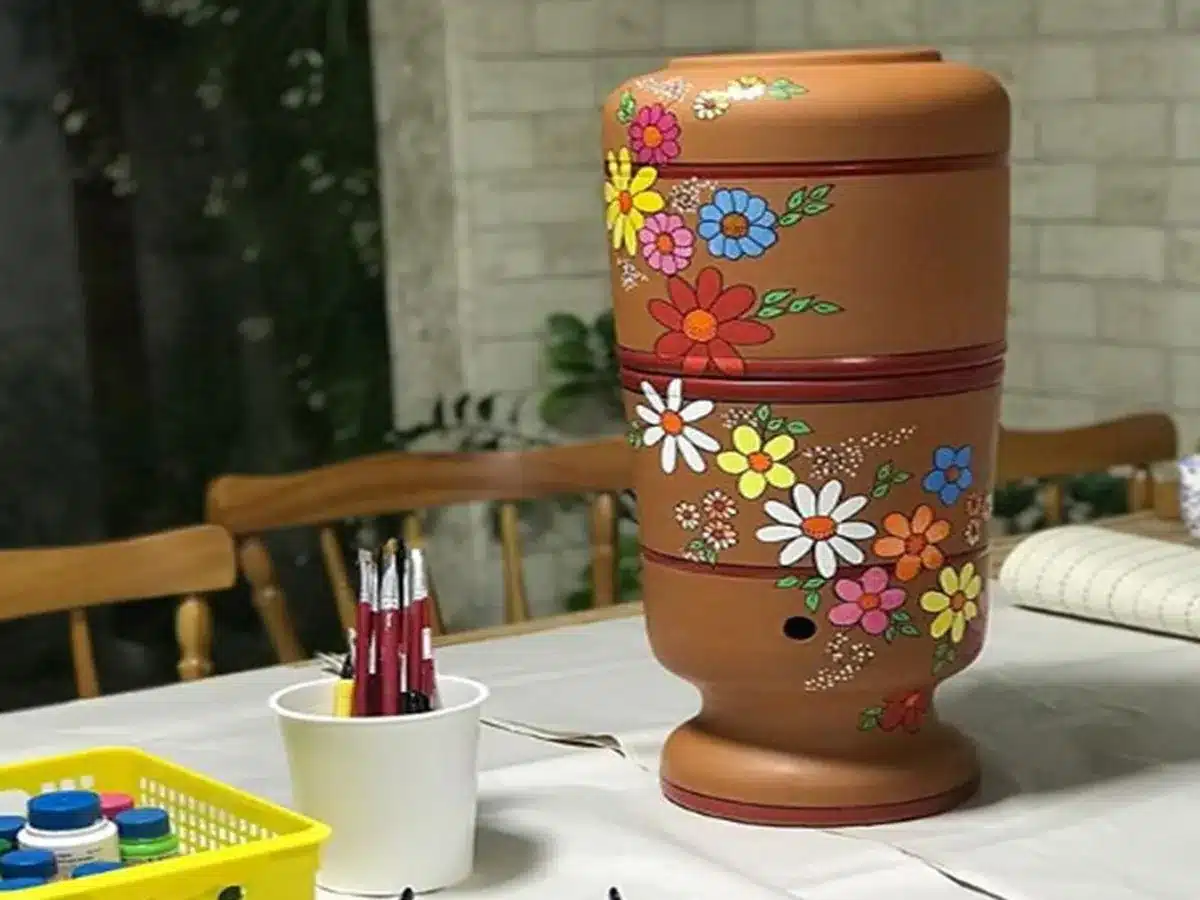 Filtro de barro decorado com flores, sobre mesa com pincéis e tintas