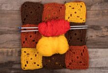 Tapete de crochê colorido composto por quadrados em tons terrosos, com novelos de linha vermelha e amarela e agulhas sobre ele, sobre fundo de madeira.