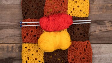 Tapete de crochê colorido composto por quadrados em tons terrosos, com novelos de linha vermelha e amarela e agulhas sobre ele, sobre fundo de madeira.