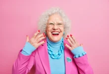 Mulher idosa sorrindo e exibindo suas unhas vermelhas, vestindo roupas coloridas em fundo rosa.