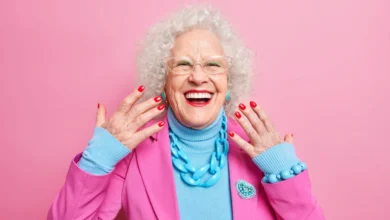 Mulher idosa sorrindo e exibindo suas unhas vermelhas, vestindo roupas coloridas em fundo rosa.
