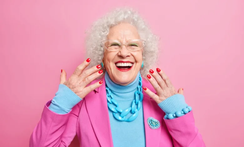 Mulher idosa sorrindo e exibindo suas unhas vermelhas, vestindo roupas coloridas em fundo rosa.