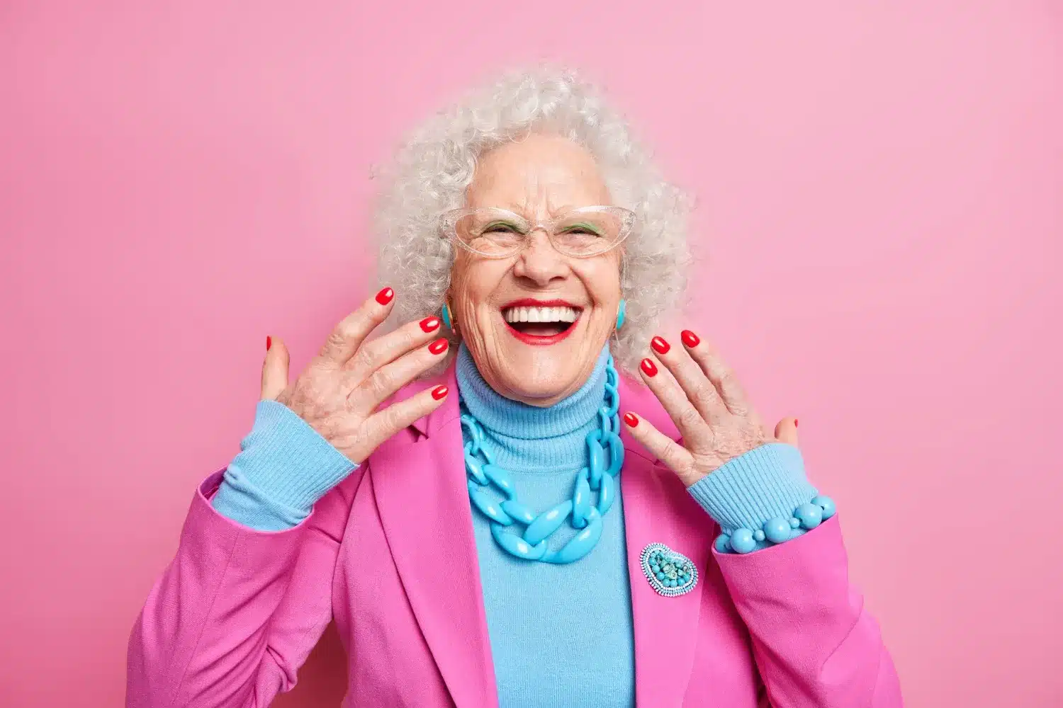Mulher idosa sorrindo e exibindo suas unhas vermelhas, vestindo roupas coloridas em fundo rosa.