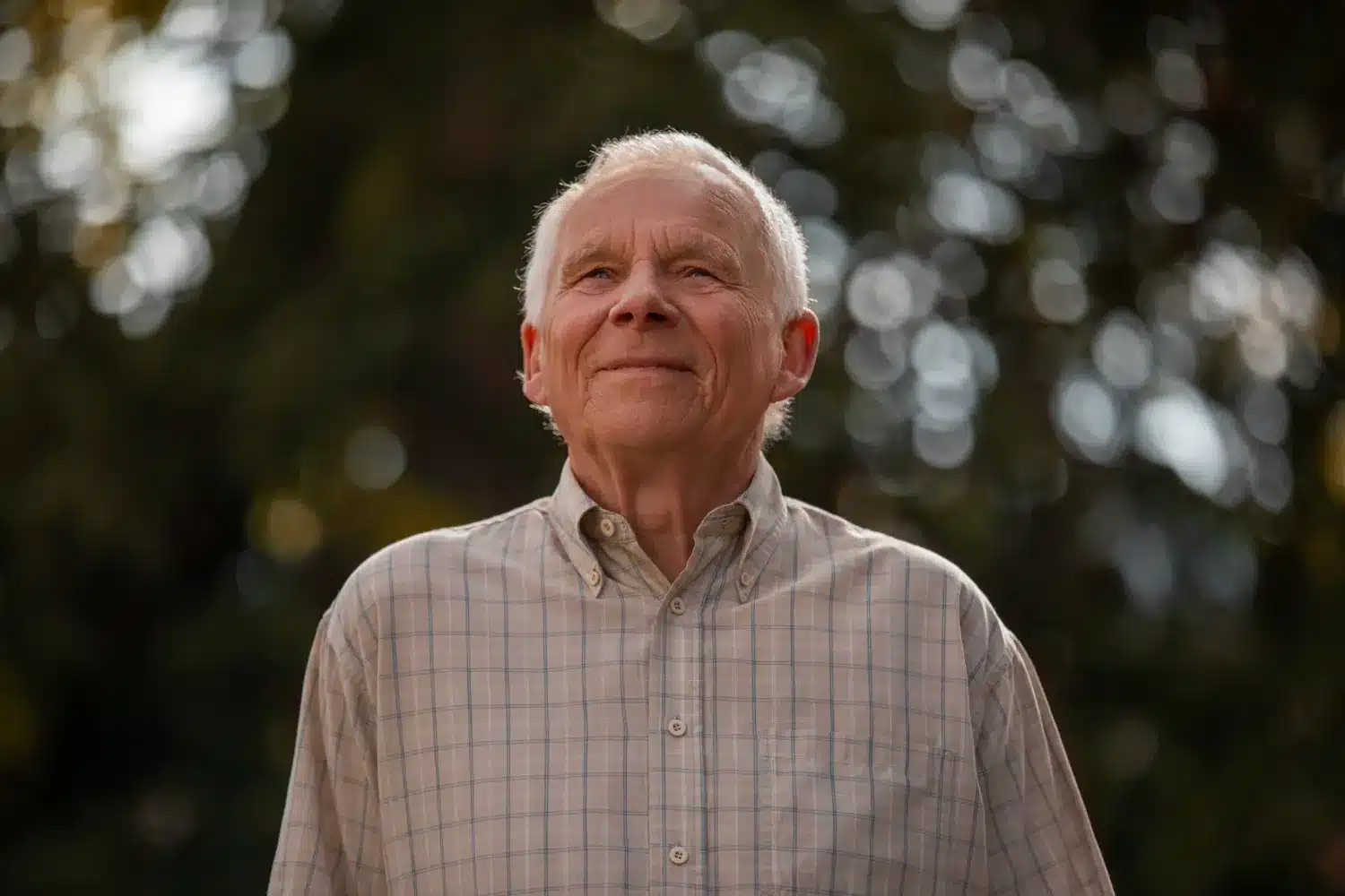 Homem idoso sorrindo, olhando para cima, em ambiente ao ar livre com luz suave