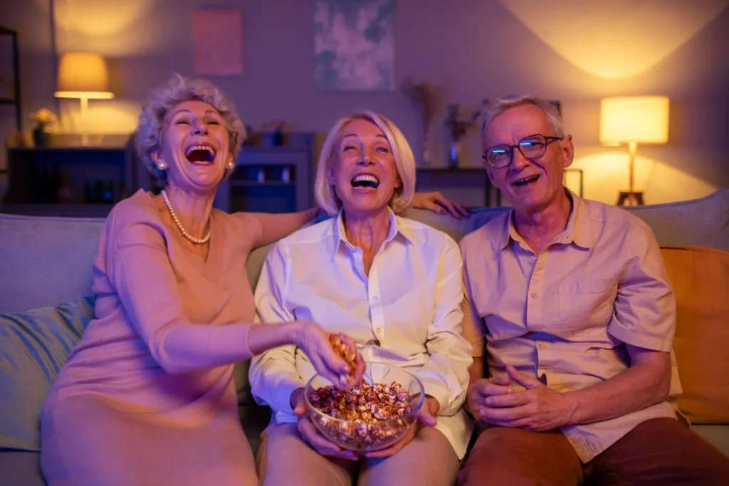 Três idosos sorrindo e se divertindo em uma festa à noite, sentados no sofá comendo pipoca.