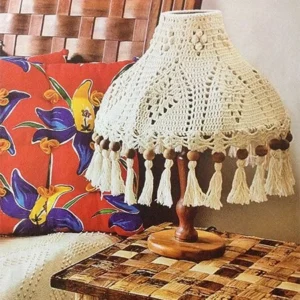 Abajur decorativo com cúpula de crochê branco e detalhes em tassel, sobre mesa de madeira