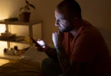 Homem sentado na cama à noite olhando para o celular, preparando-se para enviar uma mensagem de boa noite.
