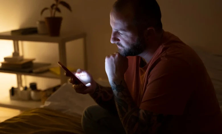 Homem sentado na cama à noite olhando para o celular, preparando-se para enviar uma mensagem de boa noite.