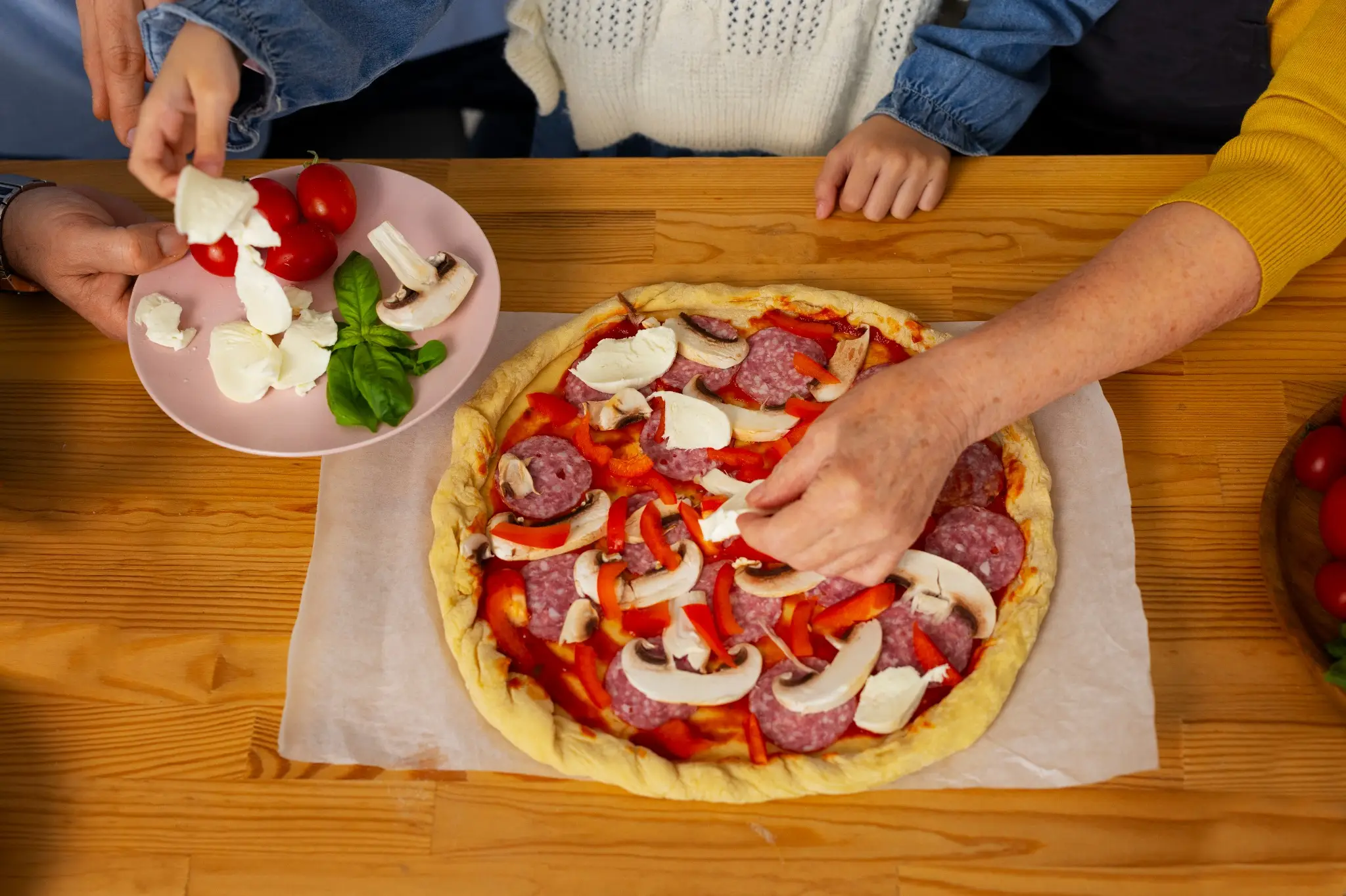 Mãos de uma criança e um adulto adicionando ingredientes à pizza caseira de pepperoni.