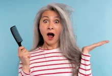 Mulher madura segurando um pente, surpresa com o cabelo bagunçado e ressecado sobre fundo azul.