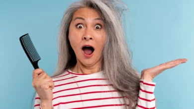 Mulher madura segurando um pente, surpresa com o cabelo bagunçado e ressecado sobre fundo azul.