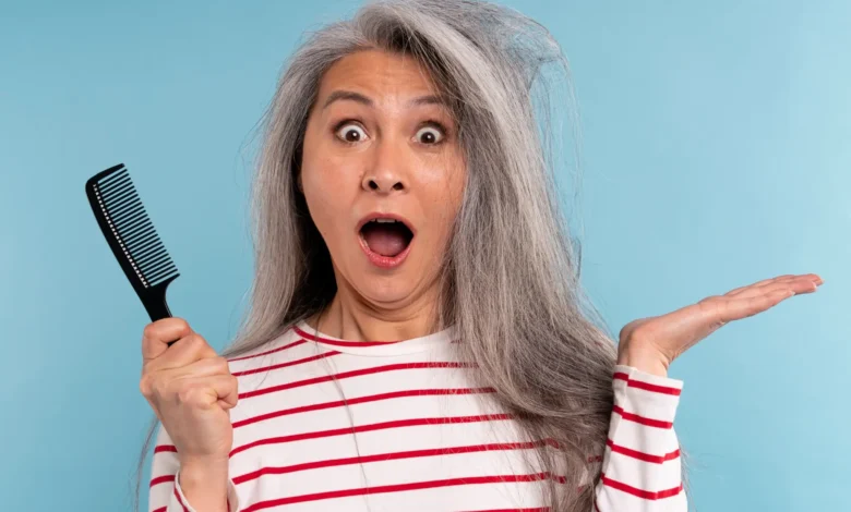 Mulher madura segurando um pente, surpresa com o cabelo bagunçado e ressecado sobre fundo azul.