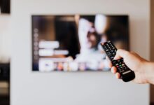 Mão segurando o controle remoto de TV enquanto assiste a uma série ou novela turca, com imagem na tela de um serviço de streaming.