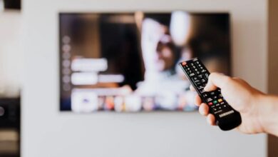 Mão segurando o controle remoto de TV enquanto assiste a uma série ou novela turca, com imagem na tela de um serviço de streaming.